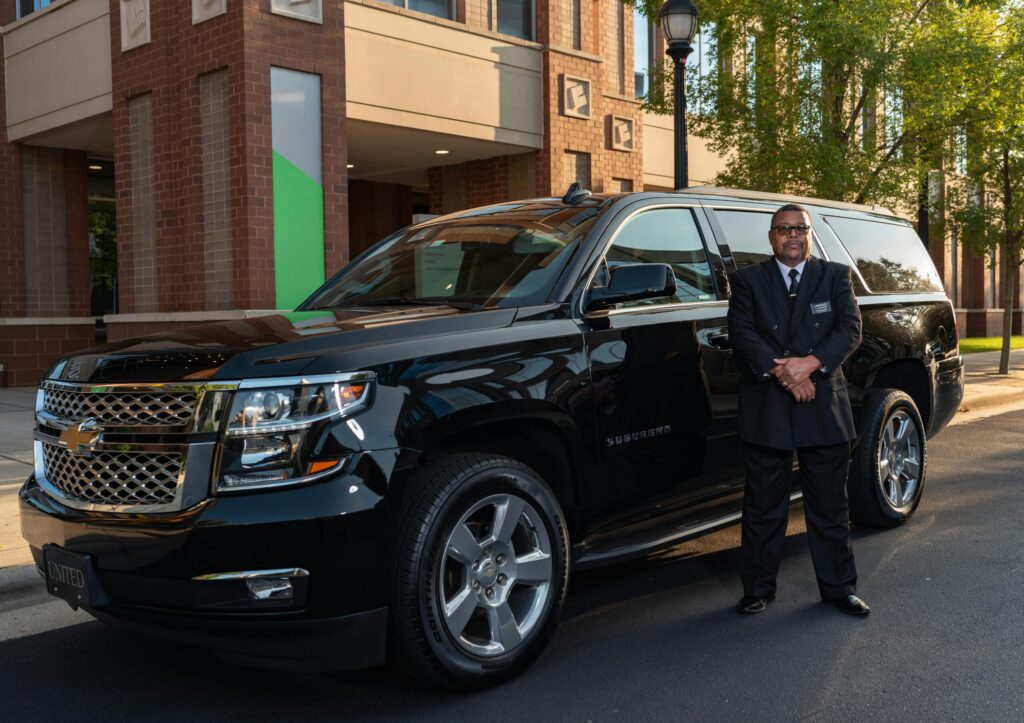 A professional chauffeur stands next to a black luxury SUV outside a modern building in daylight.
